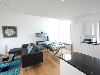 An open living and dining area with a sectional sofa, dining table with chairs, shelving unit, and kitchen cabinets at Seal Cove in Southwold