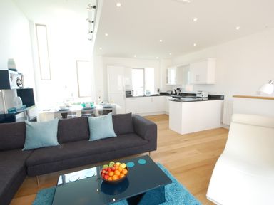 An open plan living room and kitchen with a dark sofa, blue cushions, glass coffee table with fruit bowl, white kitchen cabinets and wood floor at Seal Cove in Southwold