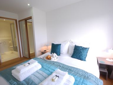 A bedroom with a bed covered in white and teal bedding a tray with croissants and a teapot bedside tables with lamps and a bathroom visible through an open door at Seal Cove in Southwold
