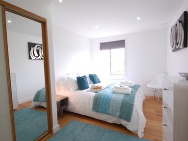 A bedroom with a double bed blue pillows and blankets a window with a blind a white chair a dresser and a mirror at Seal Cove in Southwold