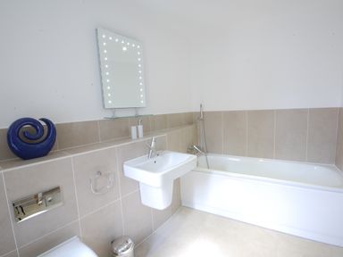 A bathroom with a bathtub, wall-mounted sink, mirror with lights, blue decorative object, and beige tiles at Seal Cove in Southwold