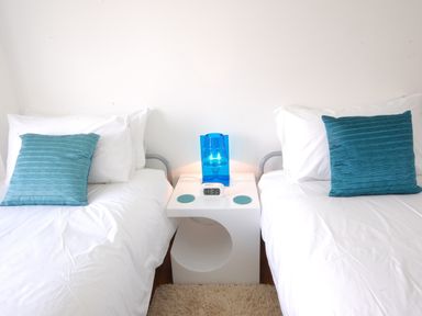 A bedroom with two single beds separated by a white bedside table with a blue lamp and a digital clock at Seal Cove in Southwold