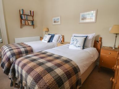 A bedroom with two single beds with checkered blankets and pillows next to wooden nightstands with lamps and a bookshelf on the wall at Crabbe House in Aldeburgh