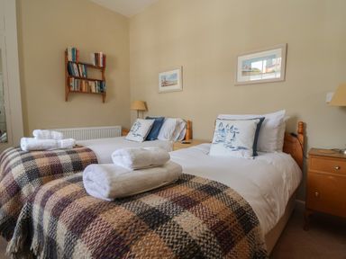 A bedroom with two single beds with plaid blankets and white towels on them a wooden bedside table with a lamp and a bookshelf on the wall at Crabbe House in Aldeburgh