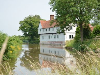 A white house with a red roof next to a calm river surrounded by trees and tall grass at The Gig House in Stowupland