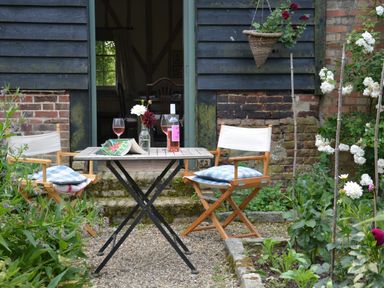 An outdoor seating area with two wooden chairs with cushions a small table with a wine bottle two glasses flowers and an open magazine in a garden at The Gig House Stowupland