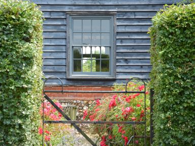 A garden with red flowers green bushes and a metal gate in front of a house with a blue wooden exterior and a gray window at The Gig House Stowupland