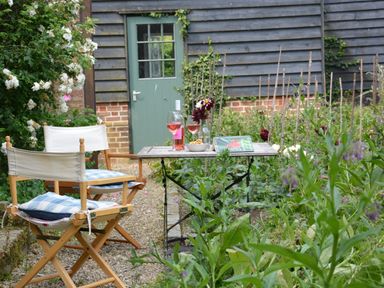A small outdoor table with two chairs beside a garden with flowers and a green door on a wooden building at The Gig House Stowupland