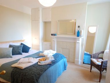 A bedroom with a bed covered with blue and gray bedding and pillows a tray with croissants and cups on the bed a white fireplace with a mirror and decorative items a wicker chair with cushions and a tall mirror near the window at Spindrift Southwold