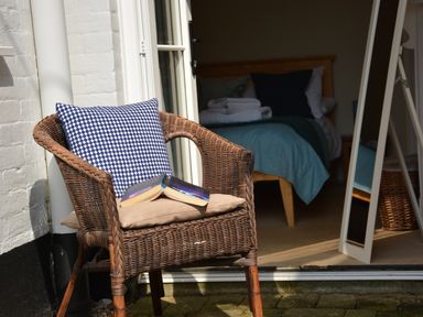 A woven chair with two cushions and an open book outside a bedroom with a bed and a mirror at Spindrift Southwold in Southwold