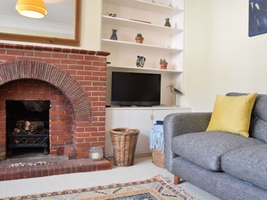 A living room with a brick fireplace a gray sofa with a yellow pillow a television on white shelves and wicker baskets at Spindrift Southwold