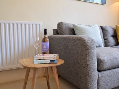 A small wooden side table with two books a bottle of wine and an empty wine glass next to a grey sofa with cushions at Spindrift Southwold in Southwold