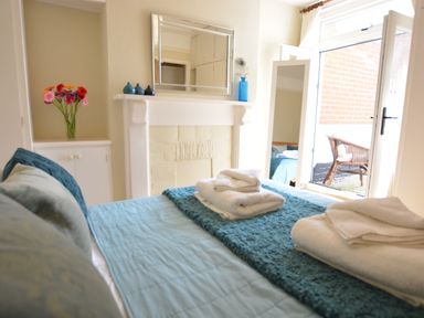 A bedroom with a bed covered with blue bedding and neatly folded towels beside a white fireplace with a mirror and vases and an open door leading to a small outdoor area with a wicker chair at Spindrift Southwold in Southwold