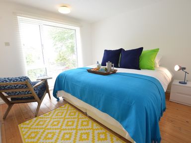 A bedroom with a bed covered in a blue blanket and pillows a blue patterned chair a yellow rug and a small white nightstand with a blue lamp at Aldeburgh Lodge Gardens in Aldeburgh