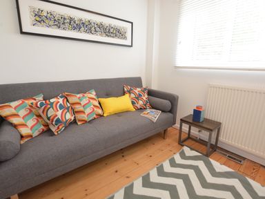 A gray sofa with colorful cushions and a small side table with a decorative item in a room with a wooden floor and a window with blinds at Aldeburgh Lodge Gardens in Aldeburgh