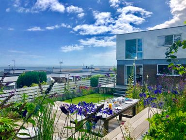 A garden with a wooden table and benches set for breakfast near a modern house with a view of the sea at Aldeburgh Lodge Gardens in Aldeburgh