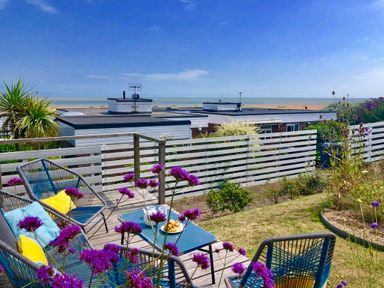 An outdoor seating area with chairs and a table on a wooden deck overlooking a garden and white fence with beach and sea in the background at Aldeburgh Lodge Gardens in Aldeburgh
