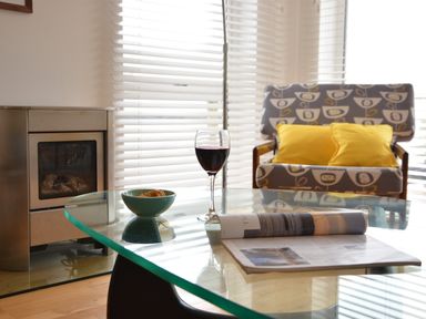 A living room with a glass table holding a bowl of snacks a glass of red wine and a magazine a patterned armchair with yellow cushions and window blinds at Aldeburgh Lodge Gardens in Aldeburgh