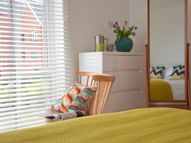 A bedroom with a wooden chair holding a decorative pillow and a magazine next to a white dresser with cups and a vase with flowers at Aldeburgh Lodge Gardens in Aldeburgh