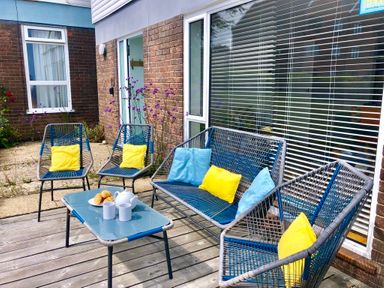 An outdoor seating area with blue chairs and sofa with yellow and blue cushions a table with a teapot two cups and croissants on a wooden deck outside Aldeburgh Lodge Gardens in Aldeburgh
