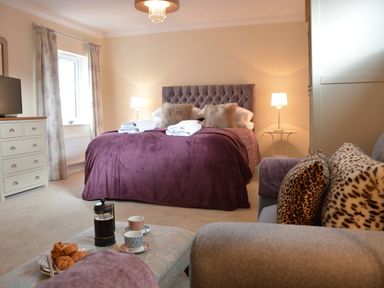 A bedroom with a bed covered in a purple blanket and two folded towels on top with two lamps on bedside tables and a sofa with animal print cushions at Meadowsweet Blythburgh in Blythburgh near Reydon