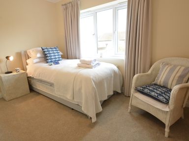 A bedroom with a single bed next to a window with beige curtains a side table with a lamp and a wicker armchair at Meadowsweet Blythburgh in Blythburgh near Reydon