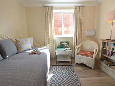 A room with a daybed with pillows and a book a small table with a Scrabble game a wicker chair a bookshelf with books and a lamp on a wooden floor with a rug at Meadowsweet Blythburgh in Blythburgh near Reydon