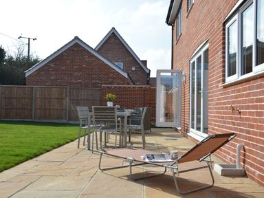 A patio with a lounge chair a dining table with chairs and a vase of flowers beside a red brick house at Meadowsweet Blythburgh