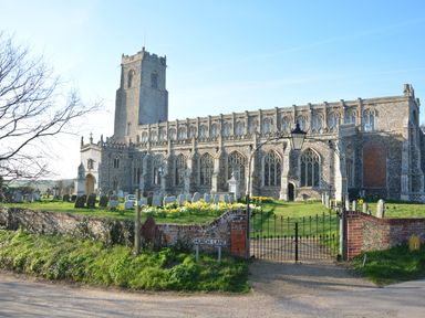 A stone church with a tower and iron gate surrounded by gravestones and flowers at Church Lane in Blythburgh near Reydon