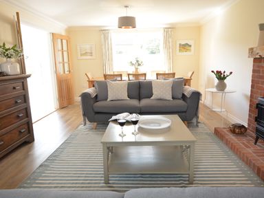 A living room with a gray sofa and coffee table with two glasses of wine and a plate with a dining table and chairs in the background at Meadowsweet, Blythburgh in Blythburgh near Reydon
