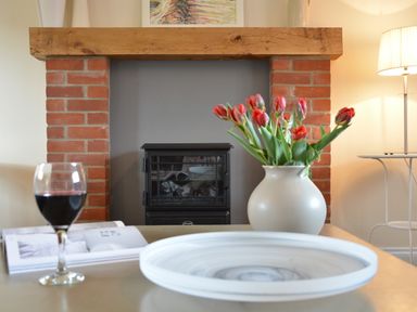 A table with a glass of red wine a book a vase with red flowers and a plate in front of a brick fireplace with a wooden mantel and a lamp on a side table