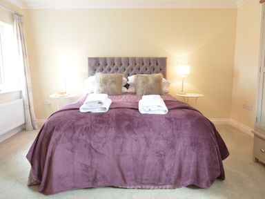 A bedroom with a bed covered in purple bedding two bedside tables with lamps and folded towels on the bed at Meadowsweet Blythburgh