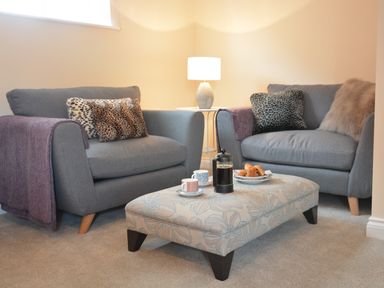A living room with two gray armchairs a floral ottoman with coffee and pastries and a lamp on a side table at Meadowsweet Blythburgh