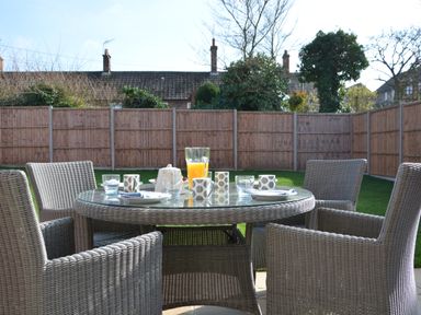 A glass outdoor dining table with wicker chairs set with cups plates and a jug in a fenced backyard at Meadowsweet Blythburgh