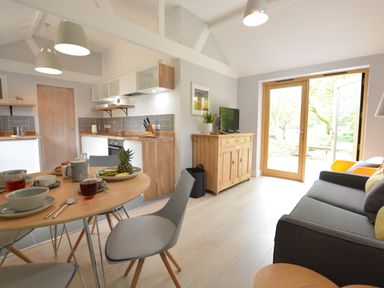A kitchen and dining area with a round wooden table set with dishes and drinks a wooden sideboard with a TV a gray sofa and glass doors opening to a garden at Milk Parlour in Spexhall Near Halesworth