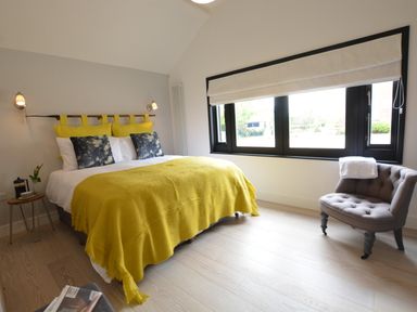 A bedroom with a double bed yellow blanket and pillows a gray chair by the window and a small wooden side table at Milk Parlour Spexhall Near Halesworth