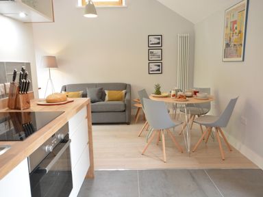 A kitchen area with a wooden counter and stove next to a dining table with gray chairs and a sofa in the background at Milk Parlour Spexhall Near Halesworth
