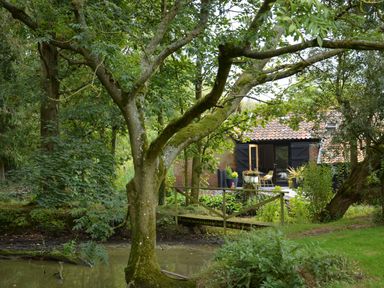 A garden with trees a small wooden bridge over a pond and a brick house with open doors at Milk Parlour Spexhall Near Halesworth