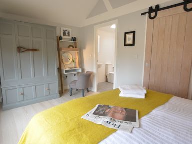 A bedroom with a bed covered by a yellow throw and folded towels a grey wardrobe a small desk with a chair and an open door leading to a bathroom at Milk Parlour in Spexhall Near Halesworth
