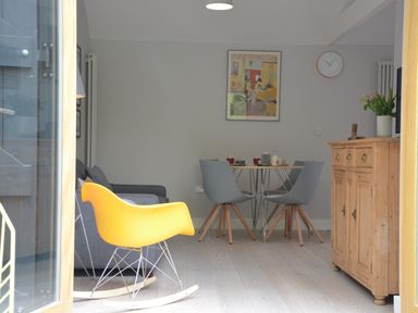A dining area with a wooden sideboard a yellow rocking chair grey chairs around a round table and a clock on the wall at Milk Parlour Spexhall Near Halesworth