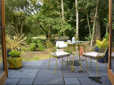 A patio with two yellow chairs and a table with a book and teacup surrounded by plants and trees at Milk Parlour Spexhall Near Halesworth