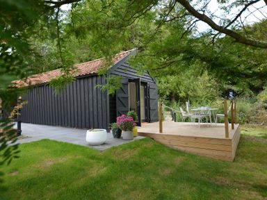 A black wooden barn with open doors next to a wooden deck with a white table and chairs in a garden at Red Poll Barn Spexhall Near Halesworth