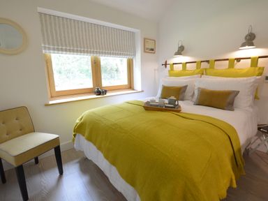 A bedroom with a bed covered in a yellow blanket and matching pillows next to a window and a chair at Red Poll Barn in Spexhall Near Halesworth