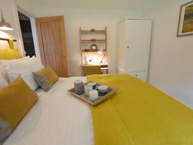 A bedroom with a bed with yellow and gray pillows a wooden tray with a tea set a wooden door a small desk with a lamp a yellow chair and a white wardrobe at Red Poll Barn Spexhall Near Halesworth