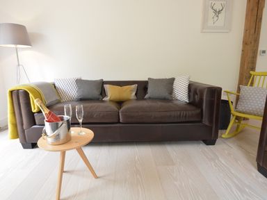 A living room with a brown leather sofa, cushions, a wooden side table with a champagne bottle and glasses, a floor lamp, and a yellow rocking chair at Red Poll Barn Spexhall Near Halesworth