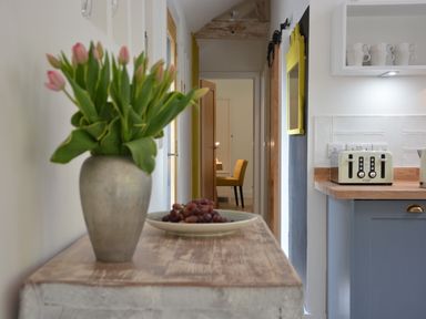 A hallway with a vase of flowers and a plate of grapes on a wooden table next to a kitchen counter with a toaster and cups at Red Poll Barn Spexhall Near Halesworth