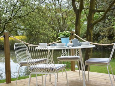 An outdoor dining table with four white metal chairs on a wooden deck near a body of water with trees around at Red Poll Barn Spexhall Near Halesworth
