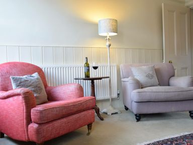 A sitting area with a red armchair and a beige loveseat with cushions a wooden side table with a wine bottle and glass and a floor lamp at Walton House Southwold