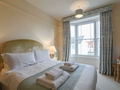 A bedroom with a bed with folded towels on it a bedside table with a lamp a window with curtains and a wooden desk with a chair at Walton House Southwold