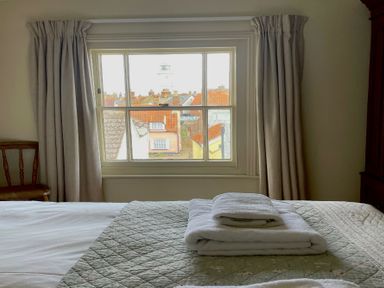 A bedroom with folded towels on a bed near a window with curtains and rooftops outside at Walton House Southwold